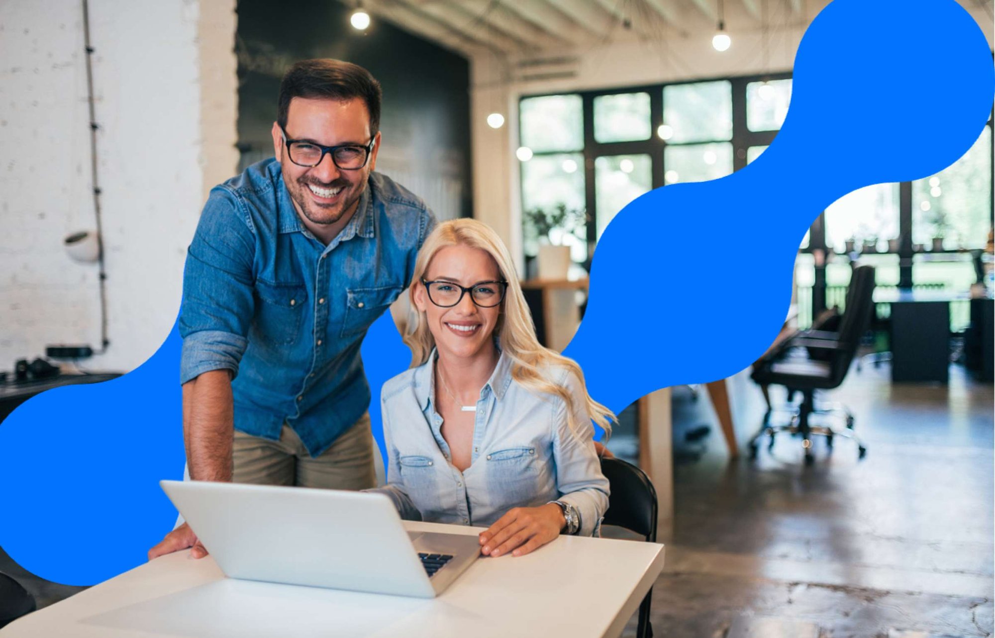 Two colleagues smiling at a laptop in a modern office, collaborating at a desk with a blue abstract background shape behind them