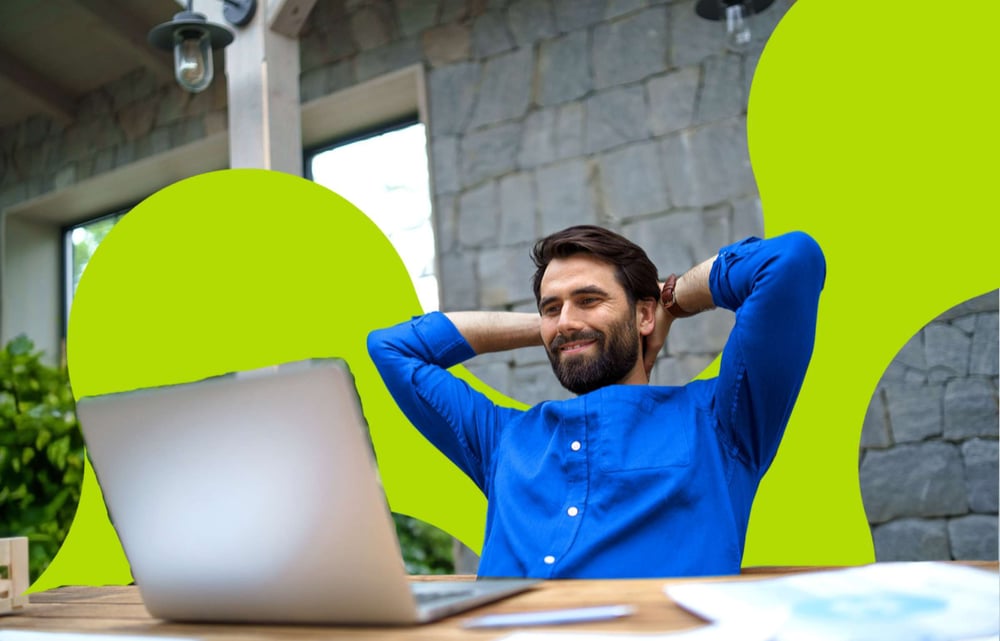 Man in a blue shirt leans back with hands behind his head, smiling at a laptop on a wooden desk