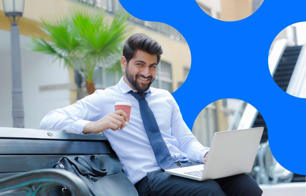 Man in business attire sitting on an outdoor bench, smiling while working on a laptop and holding a coffee cup, with a modern building and palm tree in the background