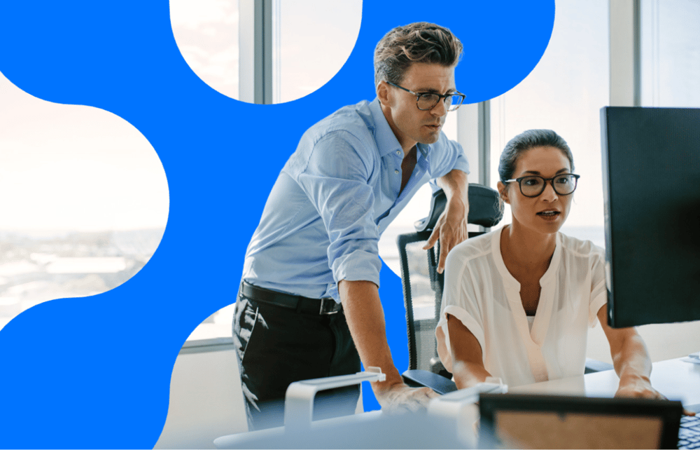 Two colleagues reviewing data on a desktop monitor in a modern office, with one standing and leaning in while the other sits and navigates